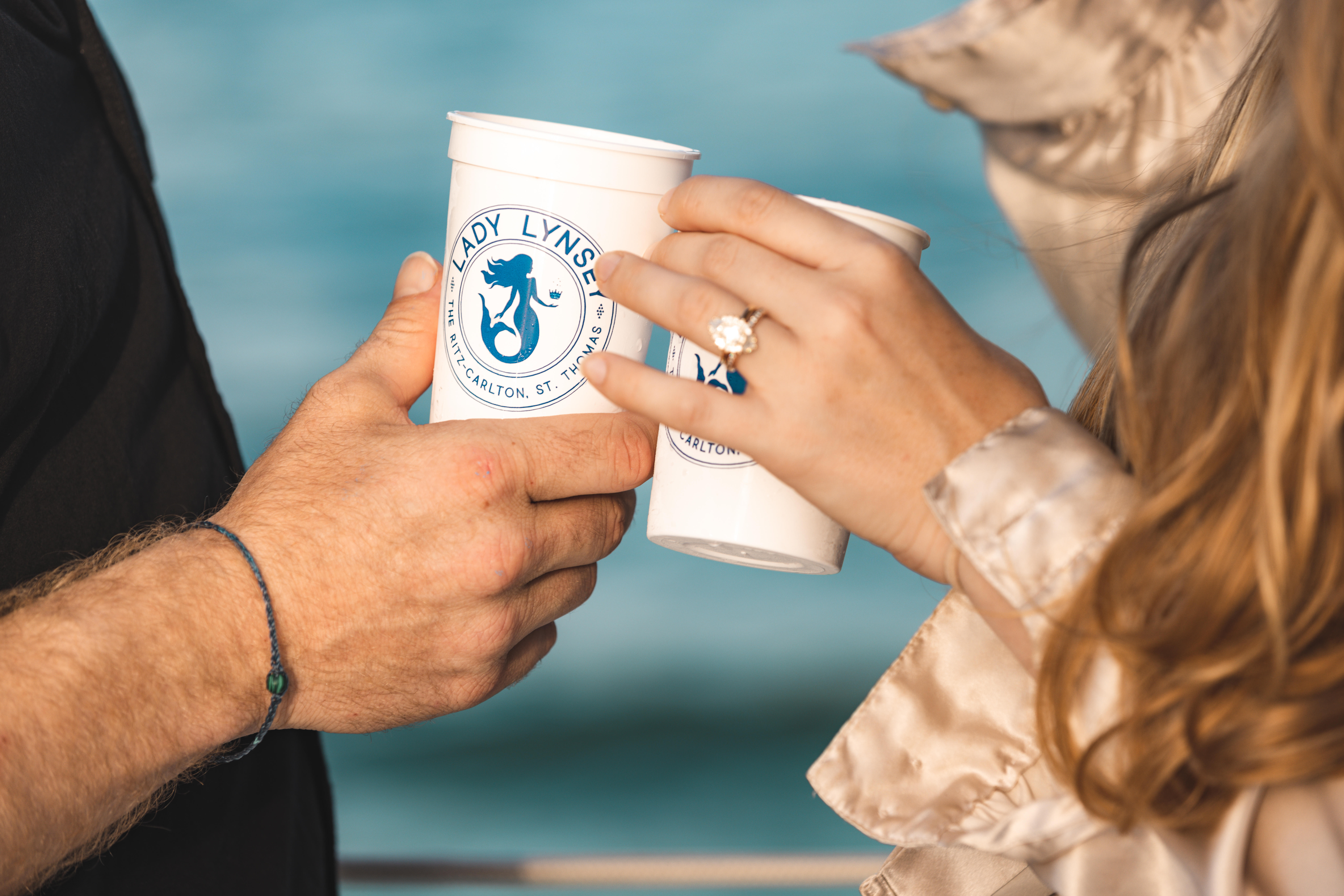 Two people putting their cups together with a blue ocean in the background