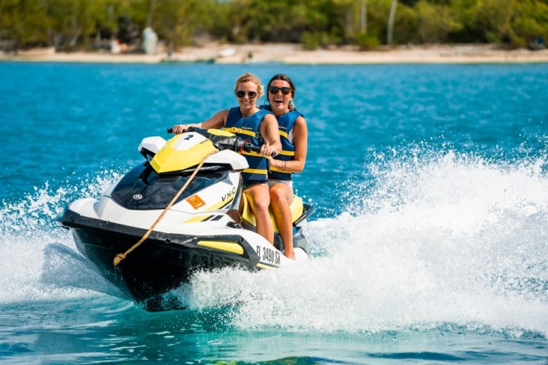 Two people riding a jet ski on clear blue water with trees in the background.