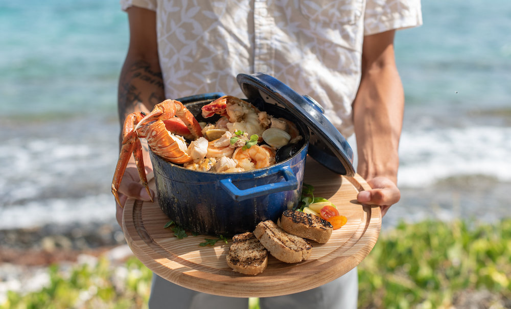Person holding seafood stew in blue pot by the ocean, with bread rolls on wooden tray.