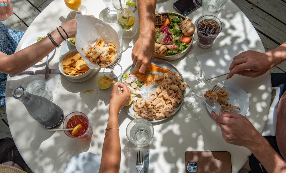 People eating salad and pasta around a sunlit table with drinks.