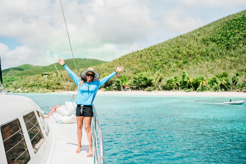 Person in blue shirt stands on boat, arms raised, with tropical island and blue water in background.