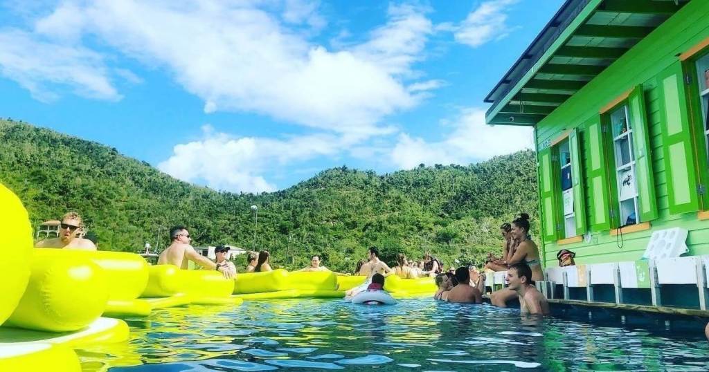 People enjoying a pool with yellow floats beside a green building, surrounded by hills under a clear sky.