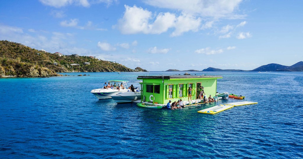 A green floating bar with people, alongside boats in clear blue water and distant islands.