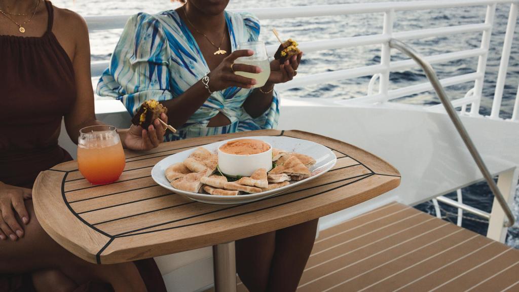 Two people on a boat enjoying drinks and snacks at a table.
