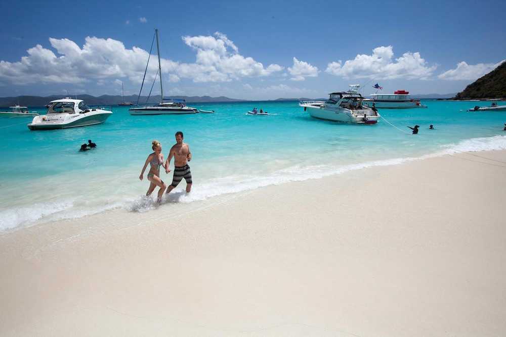 Couple walking on a sandy beach with boats in turquoise water and blue sky.