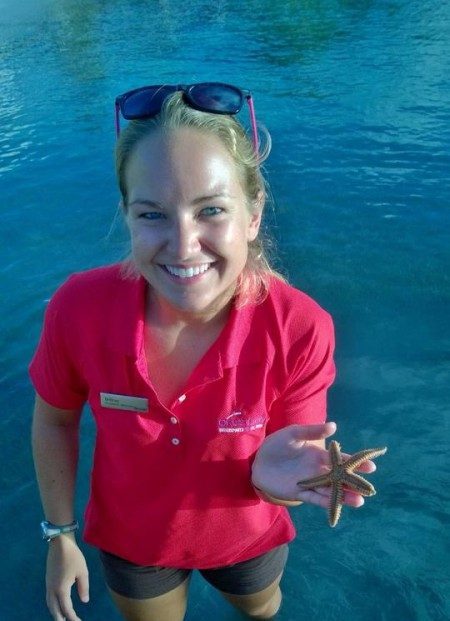 Person smiling in shallow water holding a starfish, wearing a red shirt and sunglasses.