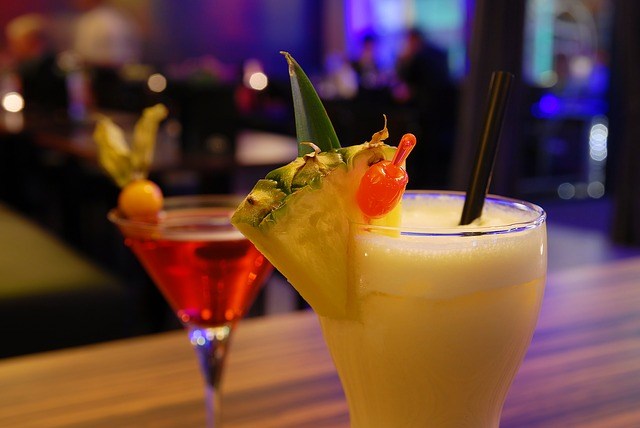 Two cocktails on a table; one with pineapple and cherry, the other with a red liquid and fruit garnish.