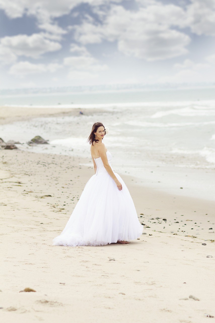 Woman in a white wedding dress walking on a sandy beach under a cloudy sky.