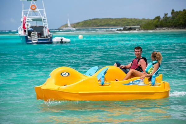 Two people enjoying a yellow paddle boat on clear turquoise water with boats and trees in the background.