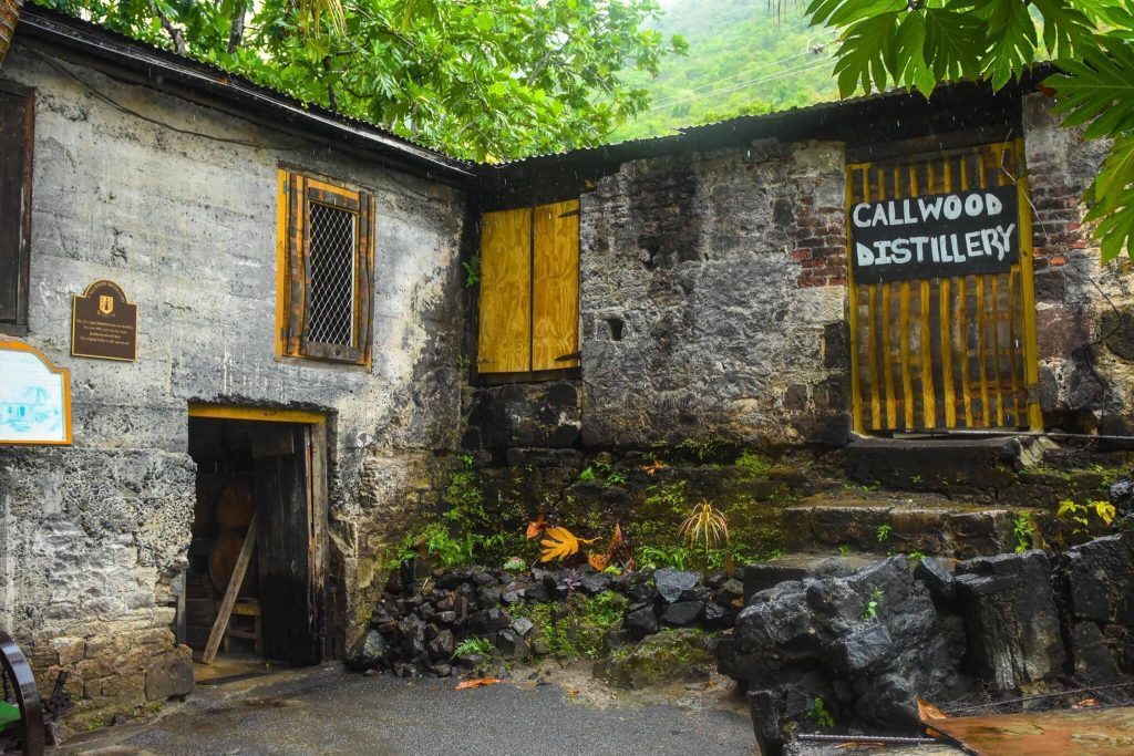 Old stone building with wooden shutters, sign reads 'Callwood Distillery,' surrounded by greenery.