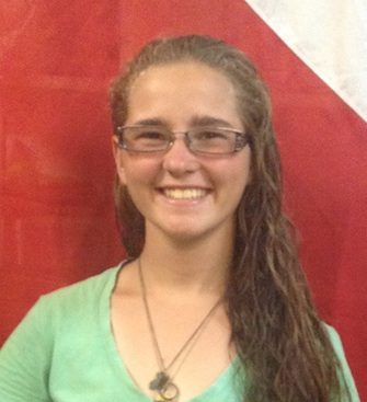 Person with glasses smiling, wearing a green shirt, standing in front of a red and white background.