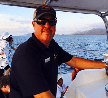 Man steering a boat on the ocean with land and sky in the background.