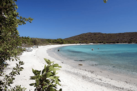 A serene beach scene with white sand, clear blue water, and a distant hill under a clear blue sky.