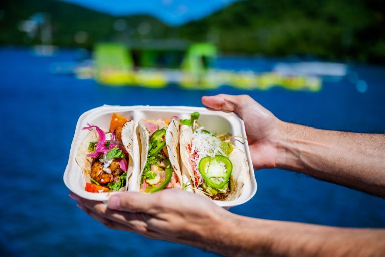 Hands holding a tray with three tacos in front of a lake view.