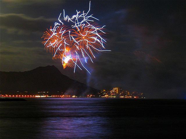 Fireworks light up the night sky over a cityscape by the sea with mountains in the background.