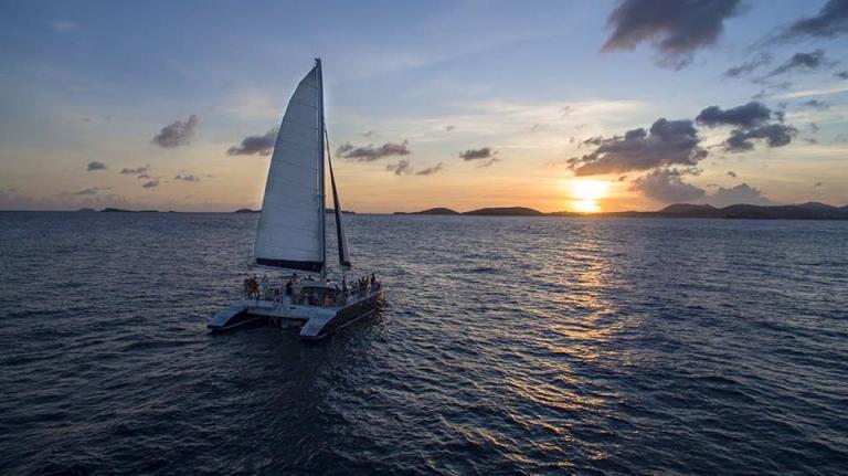 Sailboat on ocean at sunset with distant islands and clouded sky.