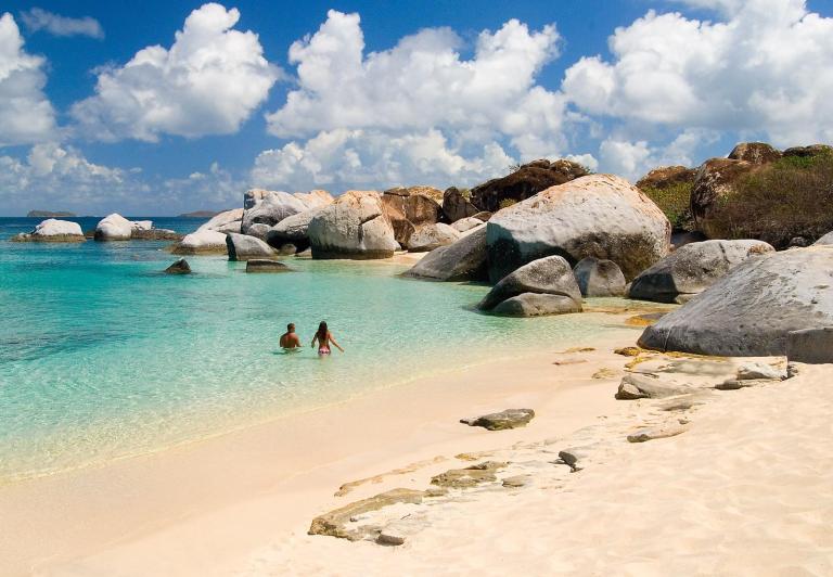 Two people wade in clear turquoise water near large rocks on a sandy beach.