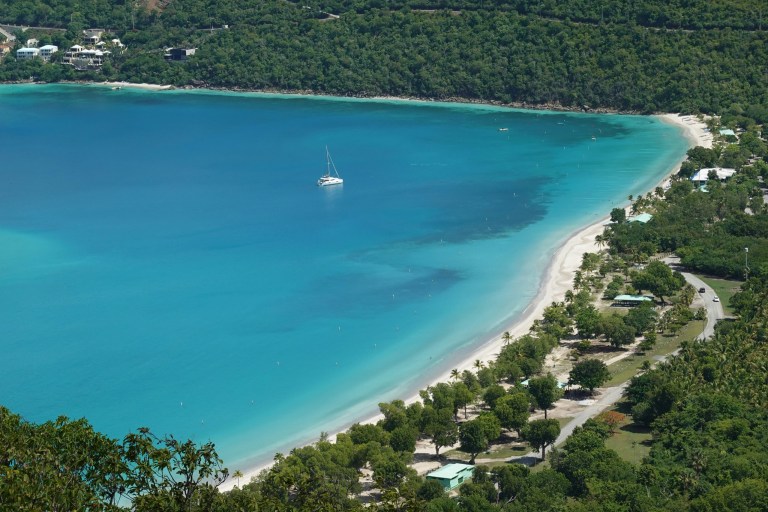 Aerial view of a sailboat on turquoise waters near a green forest and sandy beach.