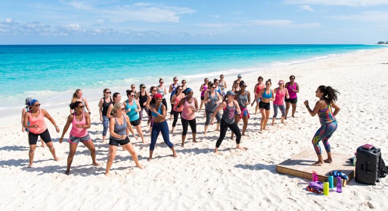 Group exercise class on beach, led by woman, ocean and blue sky in background.